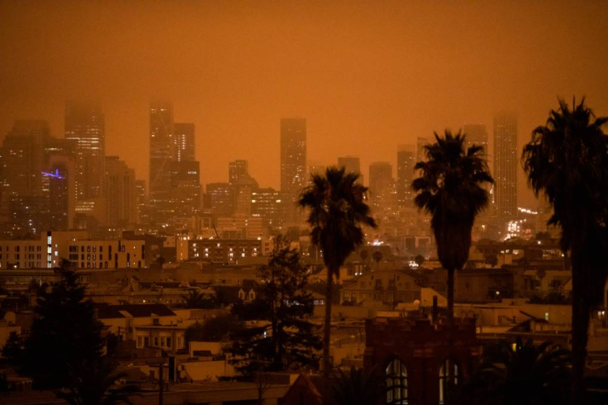Orange-tinged skies caused by smoke enshroud a city skyline. Palm trees are in the foreground