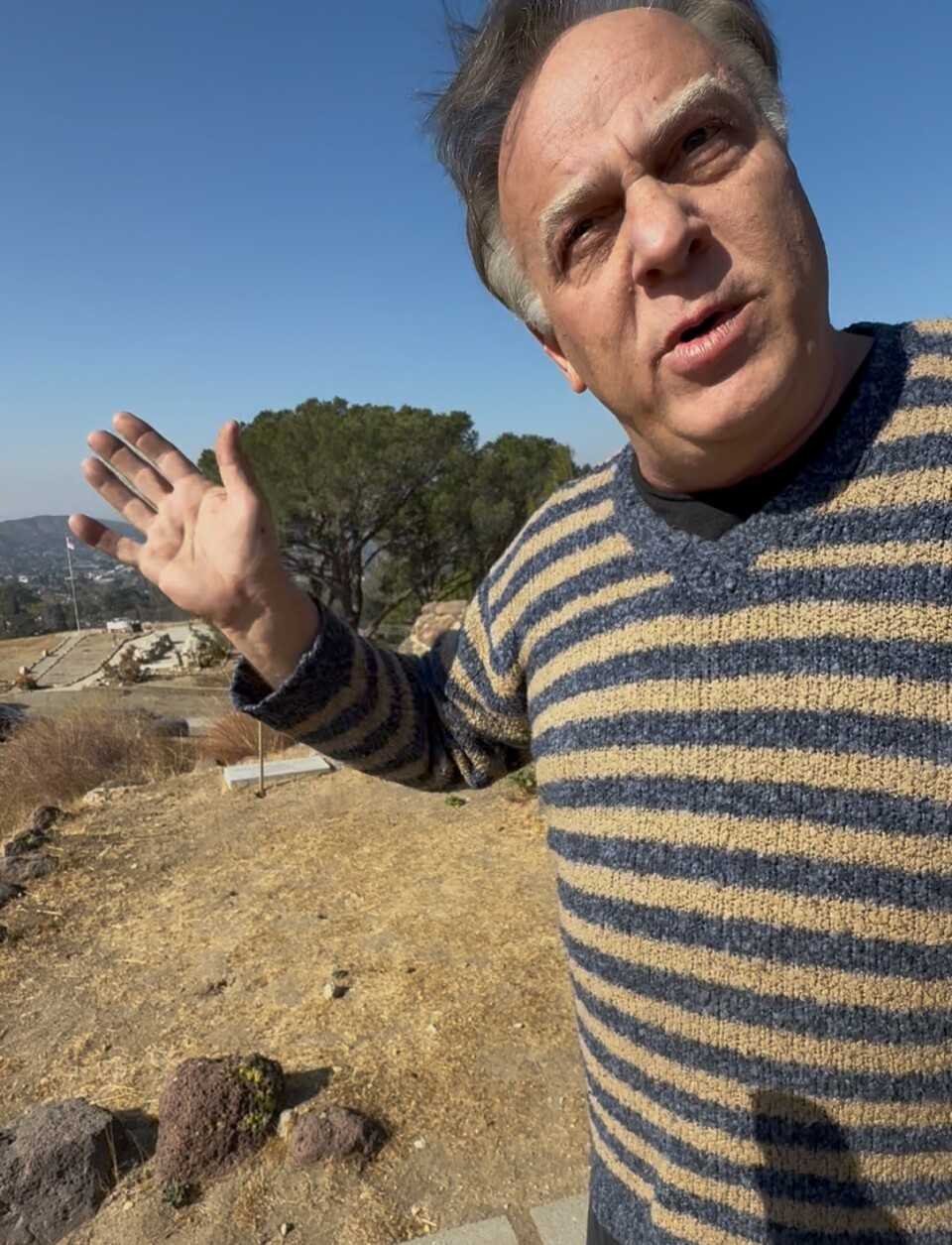 Craig Durst with Friends of Verdugo Hills Cemetery stands in the foreground. He wears a blue and white striped sweater. 