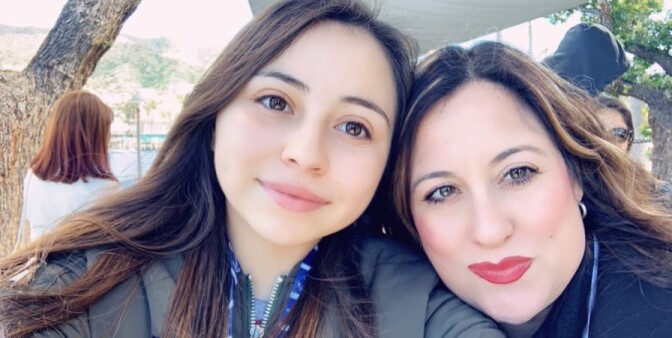 A young woman and her mother, both with long, brown hair, pose for a portrait with heads tilted and touching.