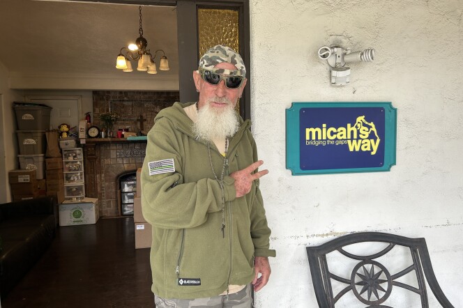 A man with a long, white beard, wearing an army green sweatshirt, sunglasses and a backwards camo-print baseball hat, stands in front of an open doorway with two fingers pointing toward an adjacent sign that reads "micah's way bridging the gaps."