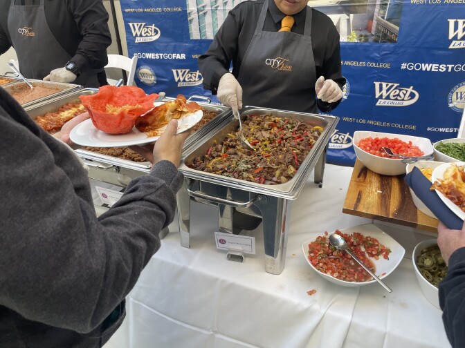A person with their back to the camera holds a plate with two hands as another person prepares to serve food from a tray. 