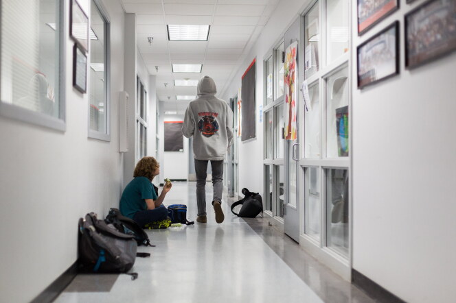 Two students in a bright school hallway: one sits on the floor against the wall eating a snack beside backpacks and a lunch bag, while another in a gray hooded sweatshirt walks away from the camera.