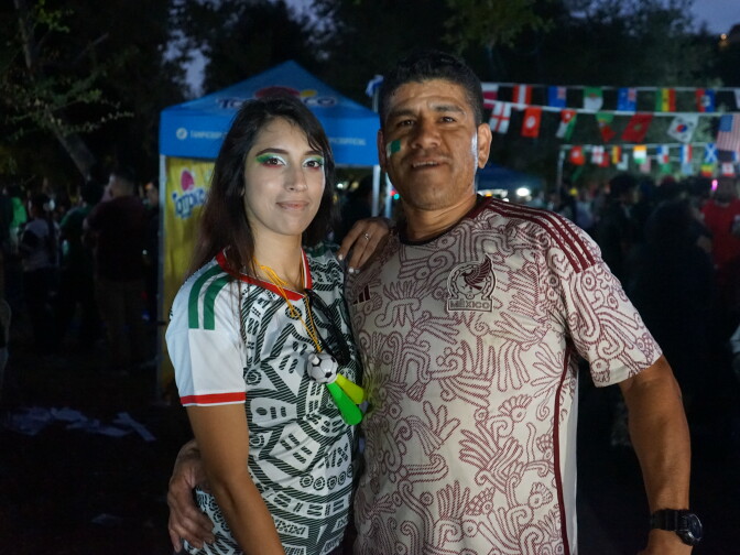 A woman and man with medium skin tones pose, arm in arm, in patterned Mexico jerseys. It's nighttime, and the surroundings are lit up by fluorescent lights. International flags are hung behind them.
