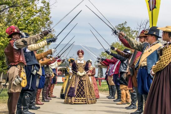 Three women dressed in voluminous Renaissance period gowns walk down an aisle outdoors, lined with men in period garb, holding their swords up and forming an awning.