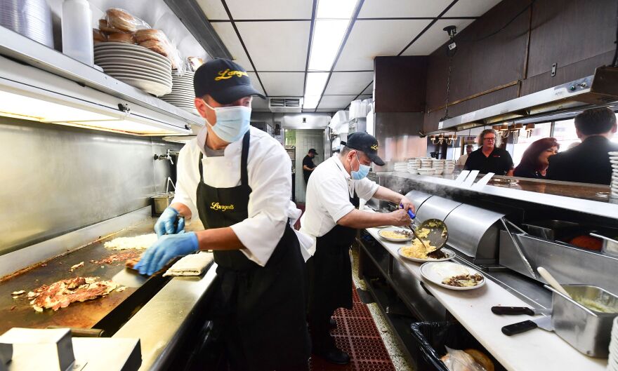 Two people with masks, aprons, and gloves cook food in a restaurant kitchen while customers wait by the counter.