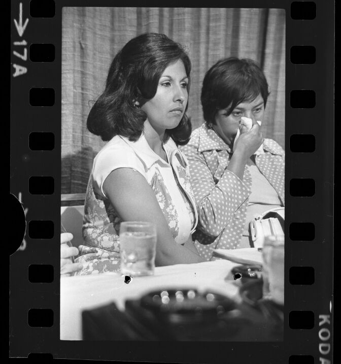 A black and white vertical photo of a Latina woman with short dark hair wearing  a button up collared dress with flower patterns. She sits very close to another older Latina woman wearing a polka dot collared long sleeve shirt and short dark hair. She holds a tissue to her nose as she blows out and looks downward. In the foreground there's a table with a glass of water and indistinguishable miscellaneous items. On the edges of the photo you can see the film perforations and the words "Kodak"