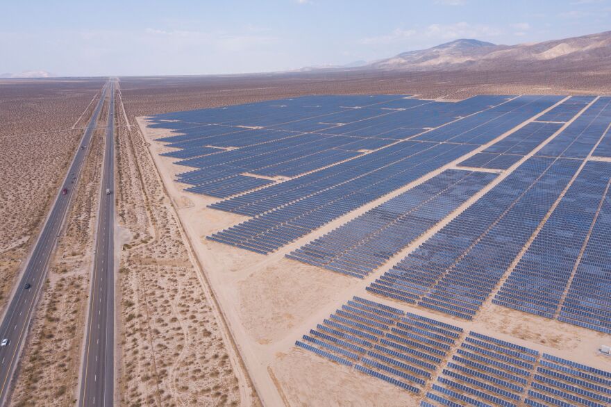 A field of solar panels is surrounded by dry flat fields with mountains in the background.