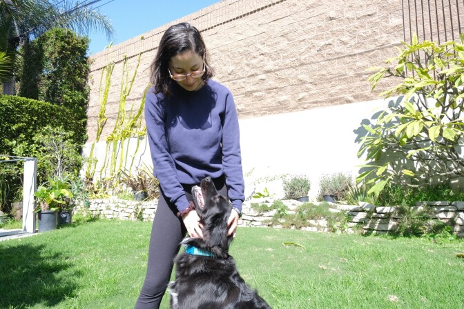 A woman with medium-tone skin and dark hair and glasses scratches a dog's neck in a backyard with a tall block freeway wall.