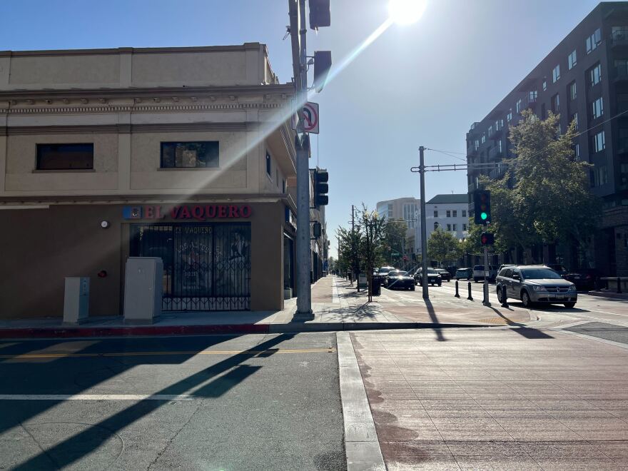 A  street corner with one empty paved street and a few cars coming on the other paved street. A ray of sunshine cuts diagonally across the image. 