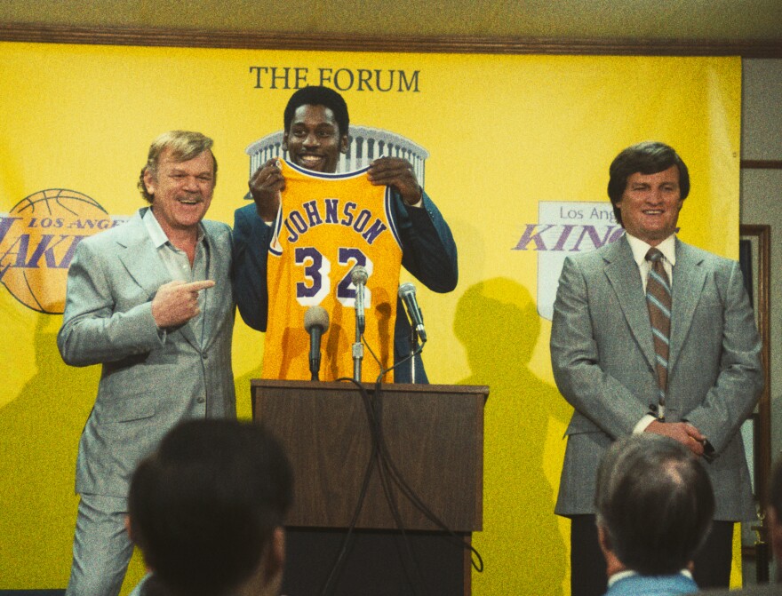 John C. Reilly on the left in a gray suit (as Dr. Jerry Buss) points to the actor portraying Magic Johnson in the middle in front of a podium with mics on it, holding up a yellow and purple Lakers jersey that reads "JOHNSON - 32." Another man in a gray suit stands on the right. They are in front of a Forum backdrop with logos for the Lakers and the Los Angeles Kings.