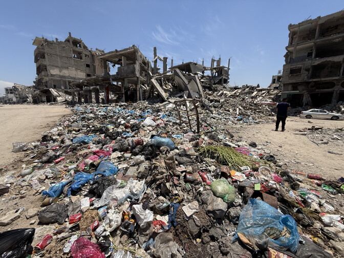 Large pile of garbage and debris with remnants of destroyed, multi-story buildings in the background.