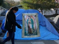 Tijuana, BAJA CALIFORNIA, Mexico - December 16, 2018. At the Benito Juarez Sports Complex near downtown Tijuana,  a woman walks past a tapestry of the Virgen de Guadalupe in the middle of the migrant tents.

 In Tijuana, Mexico, children members of the migrant caravan are learning to live in limbo as they move between shelters, settling in as much as possible to create a sense of normalcy, with help from NGOs, counselors and aid organizations. (Photo by Peggy Peattie)
