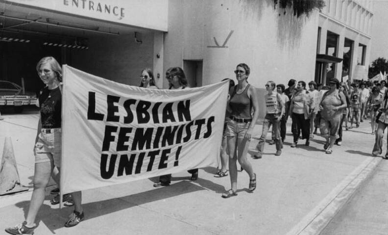A black and white photo of two people with light skin tone holding a banner that says "lesbian feminists unite." They are walking on the sidewalk ahead of a crowd of people marching.