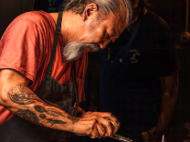 A man with a medium-dark skin tone, long gray hair in a ponytail, and a long gray goatee leans over his work at the counter, his tattooed arms visible as he prepares a dish.