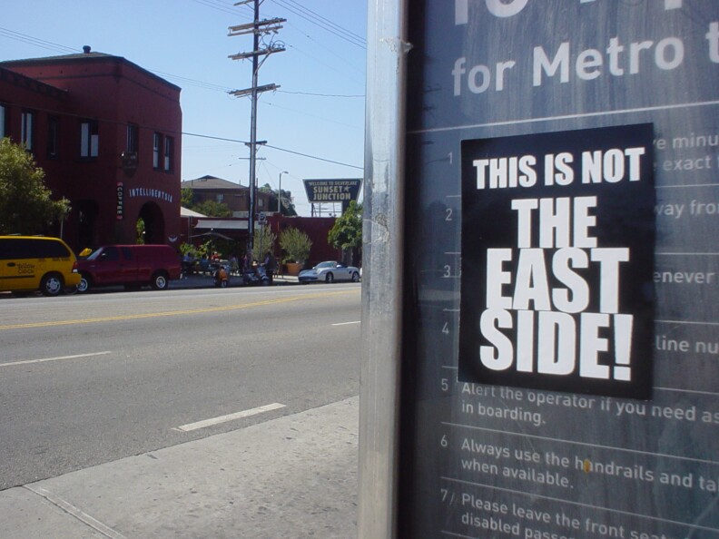 A close up of a bus stop sign that has a sticker on it. In large black and white text, it reads, "This is not the Eastside!"