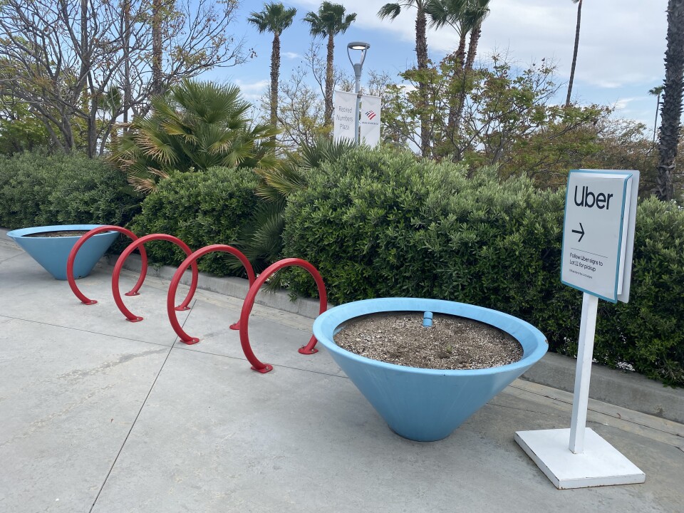 Four read bike racks are side-by-side near a large blue planter in a plaza with trees and shrubbery in the background. A sign to the right of the planter points to an Uber pickup point.