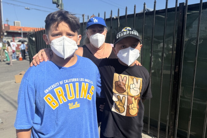 A family of three stands together in front of a black metal gate. From left to right, they include a boy wearing a blue UCLA Bruins shirt, a mother wearing a blue Dodgers baseball hat, and a younger boy wearing a black shirt featuring peace signs. All have medium skin tone and all are wearing masks. 
