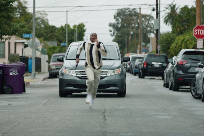 A man shows down the middle of the street as a car chases him.