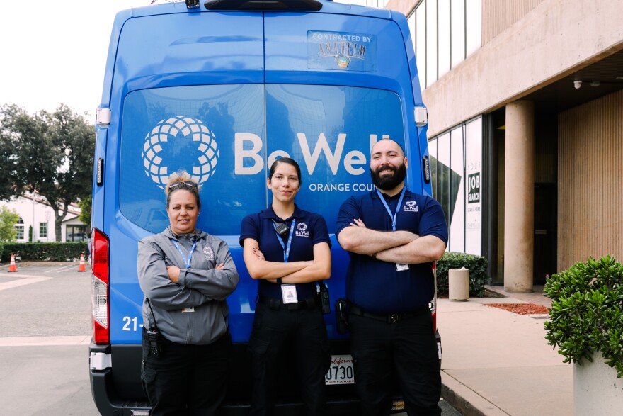 Three people stand with their arms crossed behind a blue van that read "Be Well Orange County."