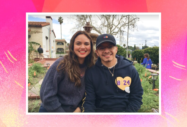 A young woman, left, and young man in dark sweatshirts sit in a garden outdoors, with greenery behind them.