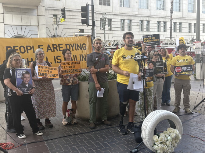 A person with a prosthetic leg speaks at a podium as others hold signs reading 'Traffic Violence Is A State Of Emergency' behind a "People Are Dying" banner.