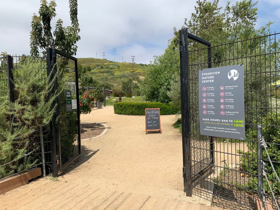 Black metal gates are propped open, inviting visitors in. The grounds are made up of dirt trails, along with manicured plots of land and raised gardening beds. There is a picnic area off into the distance, and a sign on the fence gives the park hours and rules to follow while inside. 