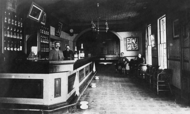 Towels hang on hooks. Two large bottles of what look like champagne sit on the bar. The bartenders have handlebar mustaches. Black and white photo.