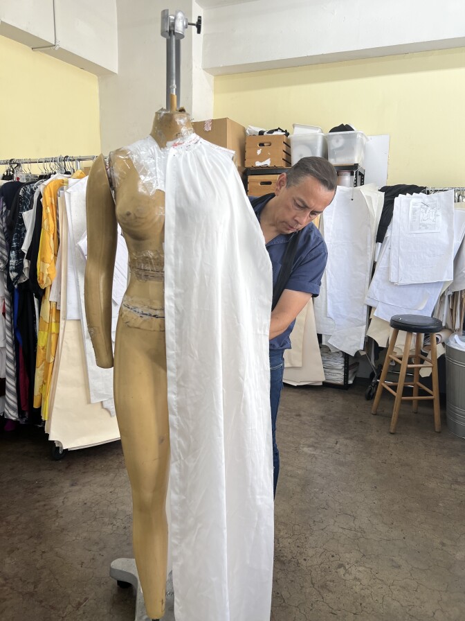 A Latino man drapes white fabric over a mannequin with racks of clothes and fabric sketches in the background