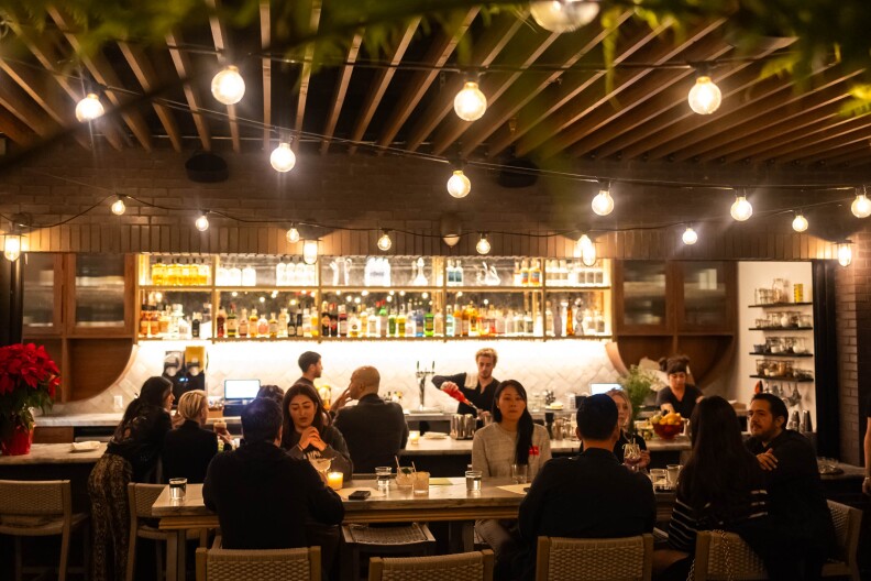 A wide photo of a the interior of a restaurant with dim lighting and a bar behind a long bar table full of people.