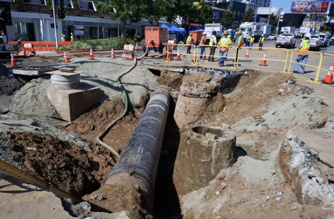 A section of a city street has been dug up, creating a deep trench with an exposed pipe running down the middle. 