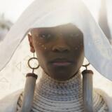 Letitia Wright stands center frame, only her shoulders up visible. She wears all white, her head hooded by a shear white fabric that falls past her shoulders. Her neck is wrapped in white beaded necklaces starting at her chest and raveling all the way up to her chin. She also wears large earrings that look like saber teeth on each ear and they too fall past her shoulders. She stares straight ahead, her face sullen. White paint is dotted into fine lines across her cheekbones and up her nose. Behind her walks a crowd of other people, also in white as if in a procession. 