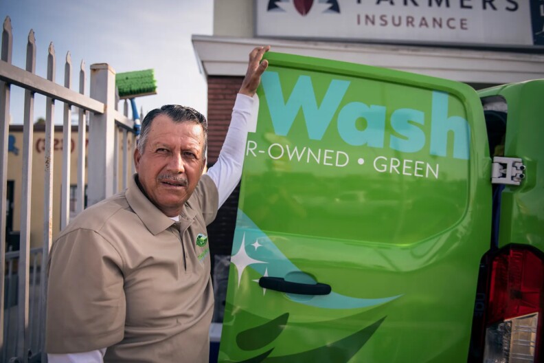 A car wash worker in a tan polo shirt poses for a photo leaning against a green car.