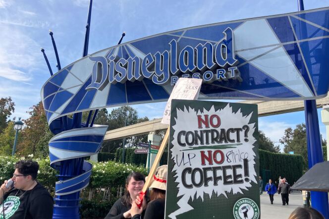 Workers stand in front of an entrance to Disneyland, which has a big sign with the park's name in blue. In front of that, a person holds a green and white sign that reads: "No Contract? No Coffee!"