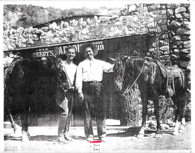 a black and white photo of two men standing between two horses and in front of a stone structure