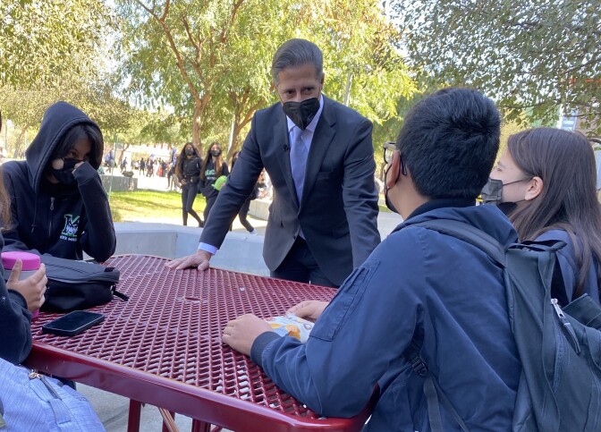 An adult wearing a suit leans over an outdoor table surrounded by middle school aged students.