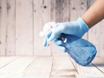 A stock image of someone with a blue glove spraying a blue bottle onto a wooden floor. 