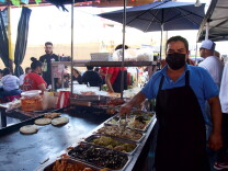 a man wearing a black apron and face mask stands next to a food cart where he is preparing tacos, taquitos and other items.