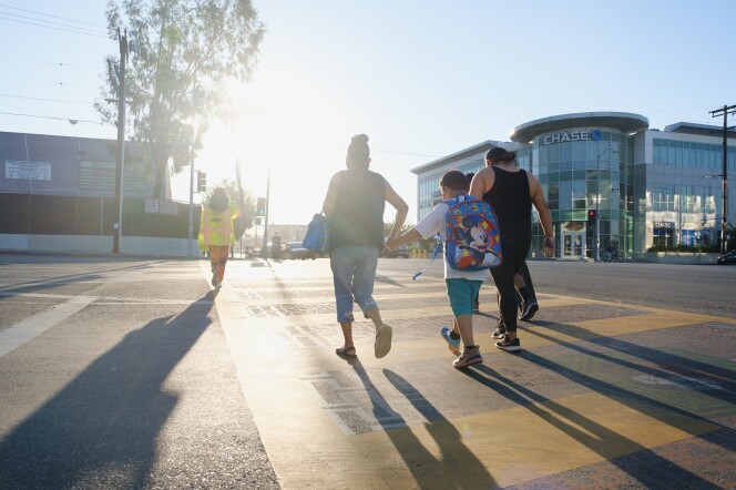 Pedestrians walk in a crosswalk at a busy intersection as a crossing guard walks ahead of them on a bright morning.