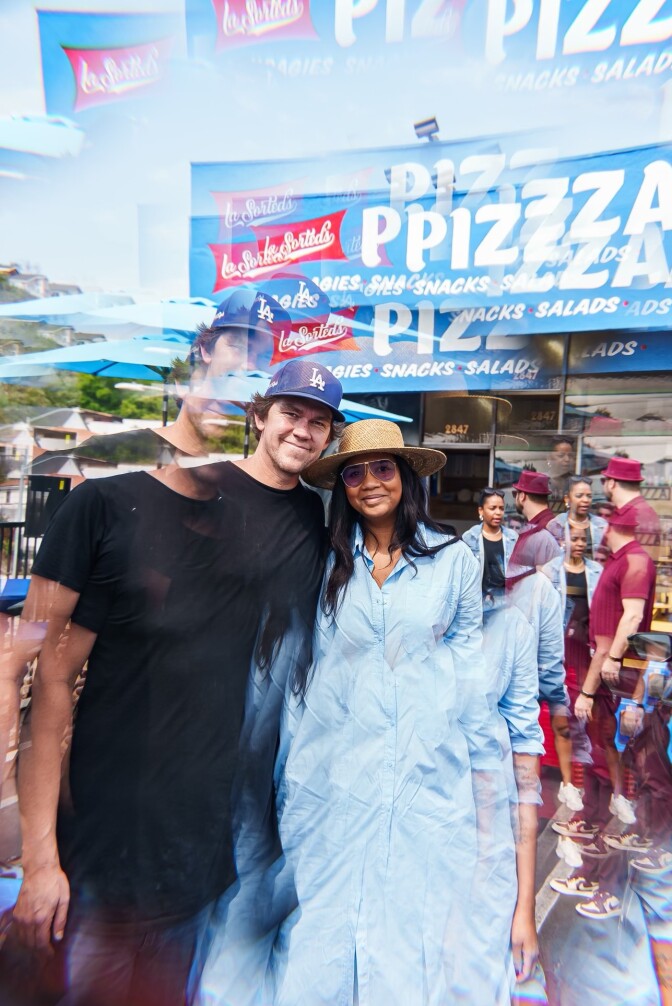 In the picture, a light-skinned toned man wearing a blue baseball cap and a black T-shirt with brown hair stands with his arms around a woman with dark skin tone and dark hair wearing a long light blue dress, a straw sun hat, and sunglasses. They are surrounded by a group of other people in the background in front of a small storefront with a light blue sign with red and white letters.
