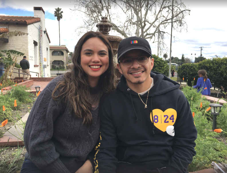 A young woman, left, and young man in dark sweatshirts sit in a garden outdoors, with greenery behind them.