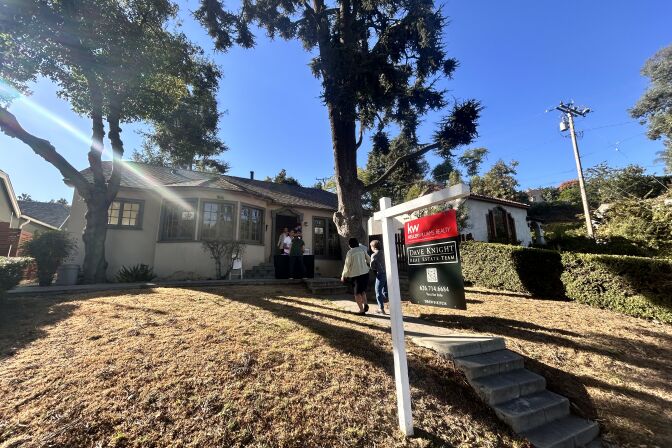 The exterior of a single-story home, with a small set of cement stairs leading towards the front door. A for sale sign is sticking out of the dried out grass on a white post.