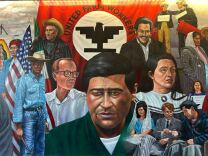A wide view of a large, ceiling to floor mural inside a college boulding. It depicts multiple labor leaders, including Dolores Huerta, surrounding Chavez in the center. In the background is the United Farm Workers union flag, which is red, with a black eagle symbol in the middle of a white cirlce.