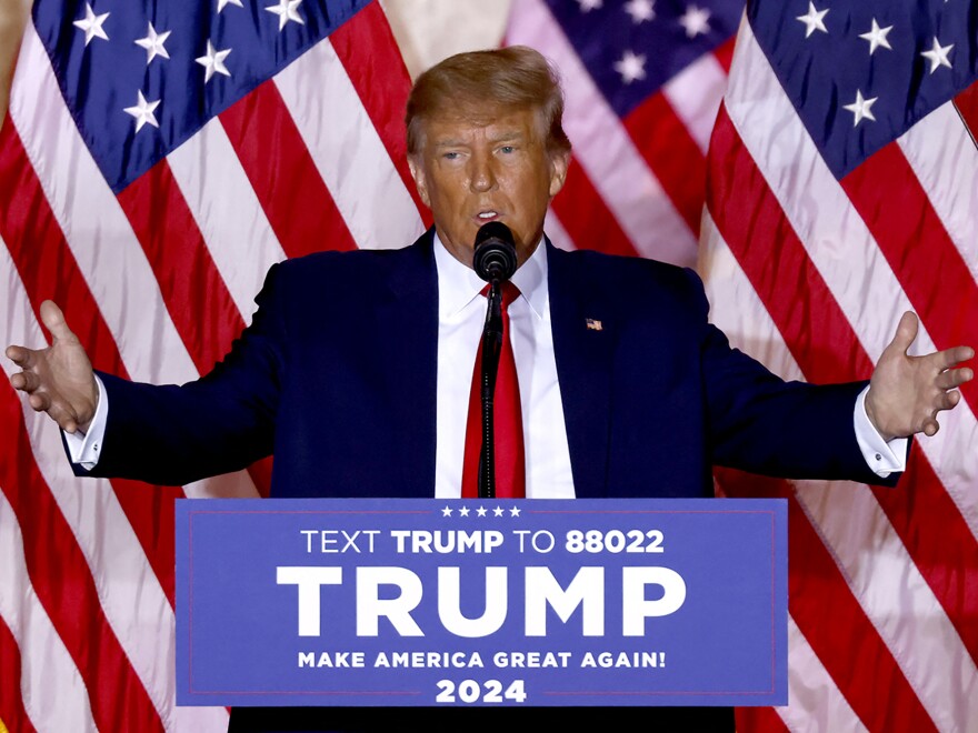 Donald Trump at a lectern with his name on it and the slogan "Make America Great Again!" Three U.S. flags are visible behind him.