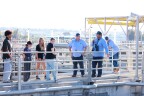 A group of high school students hear from adult water professionals in light blue attire at a water treatment facility outside on a sunny day. 