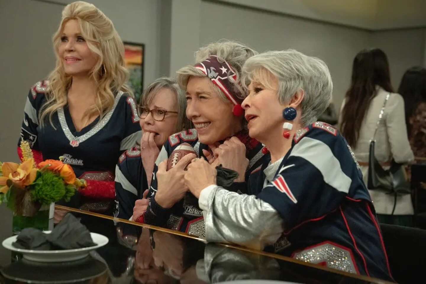 Rita Moreno, Sally Field, Lily Tomlin, and Jane Fonda sit at bar counter in a reception hall. Moreno stands while the other women sit. They are all watching something out of frame, each wearing bedazzled Patriots jerseys with various red, white, and blue accessories. Moreno smiles softly. Field bits her nails on her left hand while gripping the arm of Tomlin with her right. Tomlin holds a miniature football in her left hand and holds hands with Fonda with her left. Tomlin smiles wide, excited. Fonda looks on as if infatuated. 