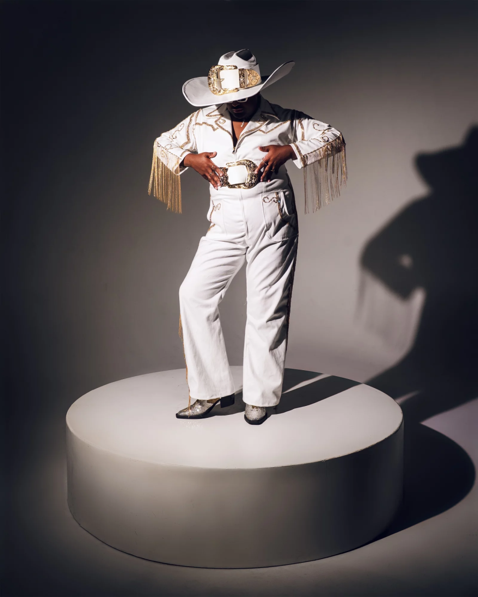 A man with dark-tone skin poses on a white platform in white jumpsuit with gold fringe and an oversized cowboy hat.