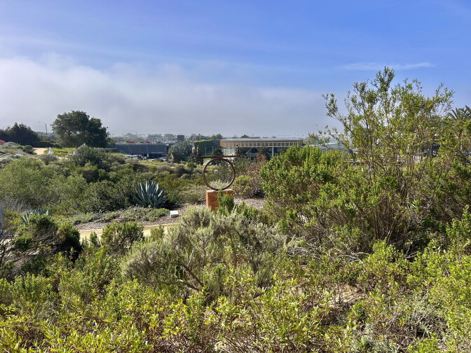 A circular metal sculpture stands amid native plants and shrubs, with a building and parking lot visible in the hazy distance.