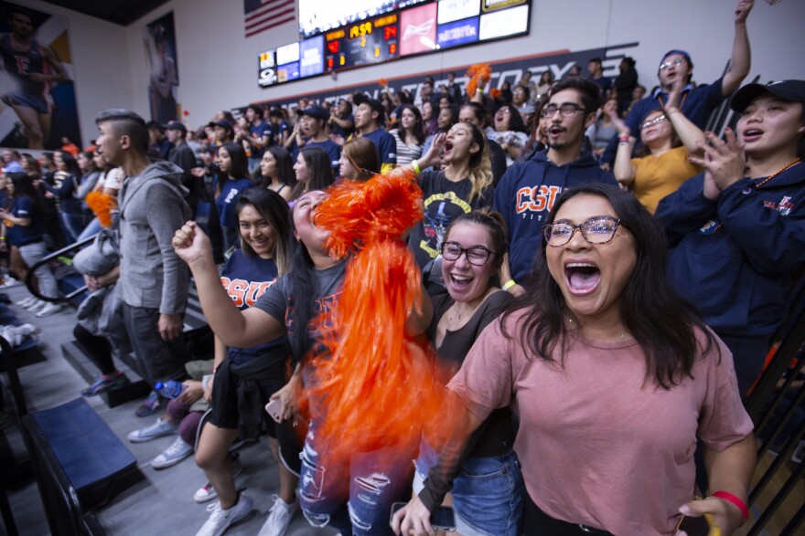 Excited fans dressed in orange and blue cheer and wave pom-poms while standing on stadium bleachers.