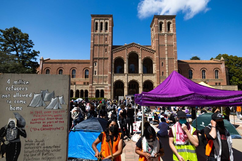Crowds on a lawn in front of a large brick university building. In the foreground to the left of frame there's a large wooden painting of tents and a Palestinian woman carrying items with text that reads "4 million refugees not allowed to come home " "in 1948 over 750,000 Palestinians were expelled from their homes as part of the Nakba "The catastrophe." While Jewish peoples of all nations are guaranteed Israeli citizenship under the law of return."