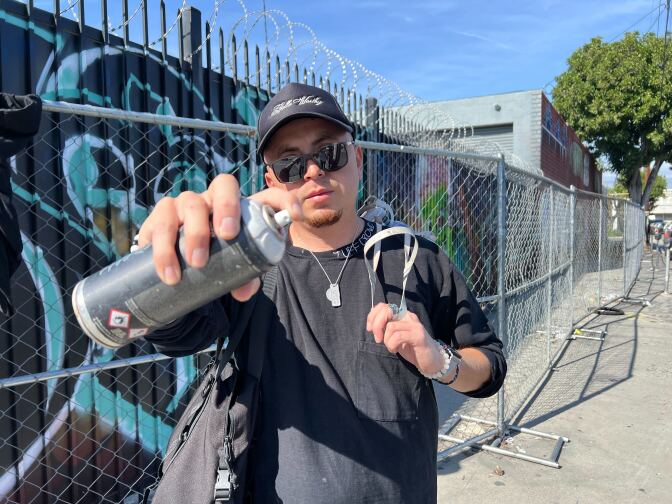 A light skin Latino man holds a spray can to the camera. In the background, there's a chainlink fence and behind that there's a wall with some graffiti art.